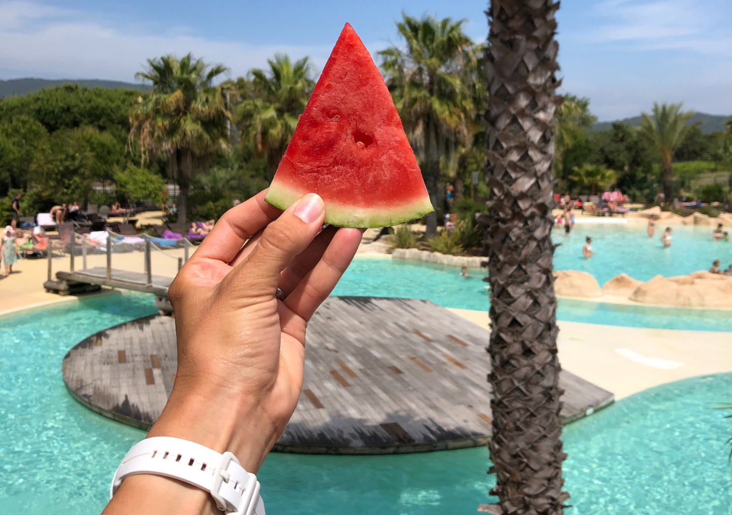 A woman's hand holds a watermelon in the background you can see the swimming pools of the Les Tournels campsite in France